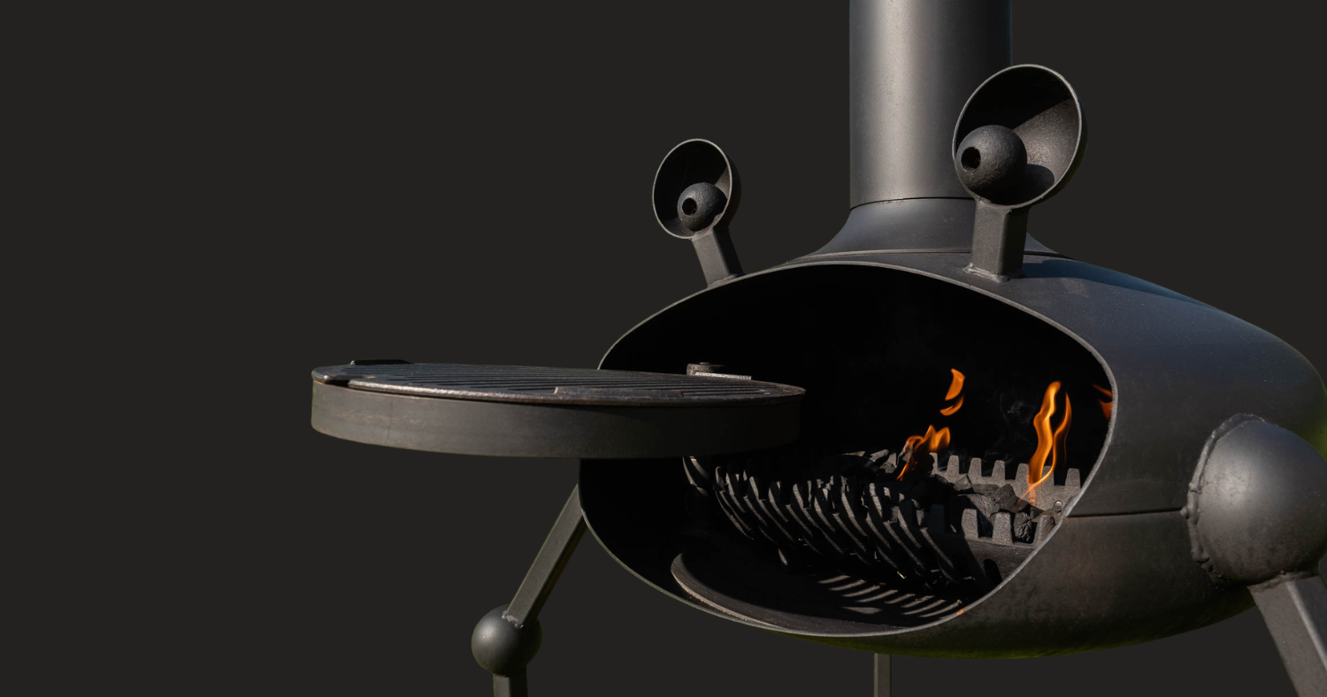 A close-up of a dark grey beast style fireplace with a circular door swung open, revealing glowing orange flames inside, set against a solid dark grey background.