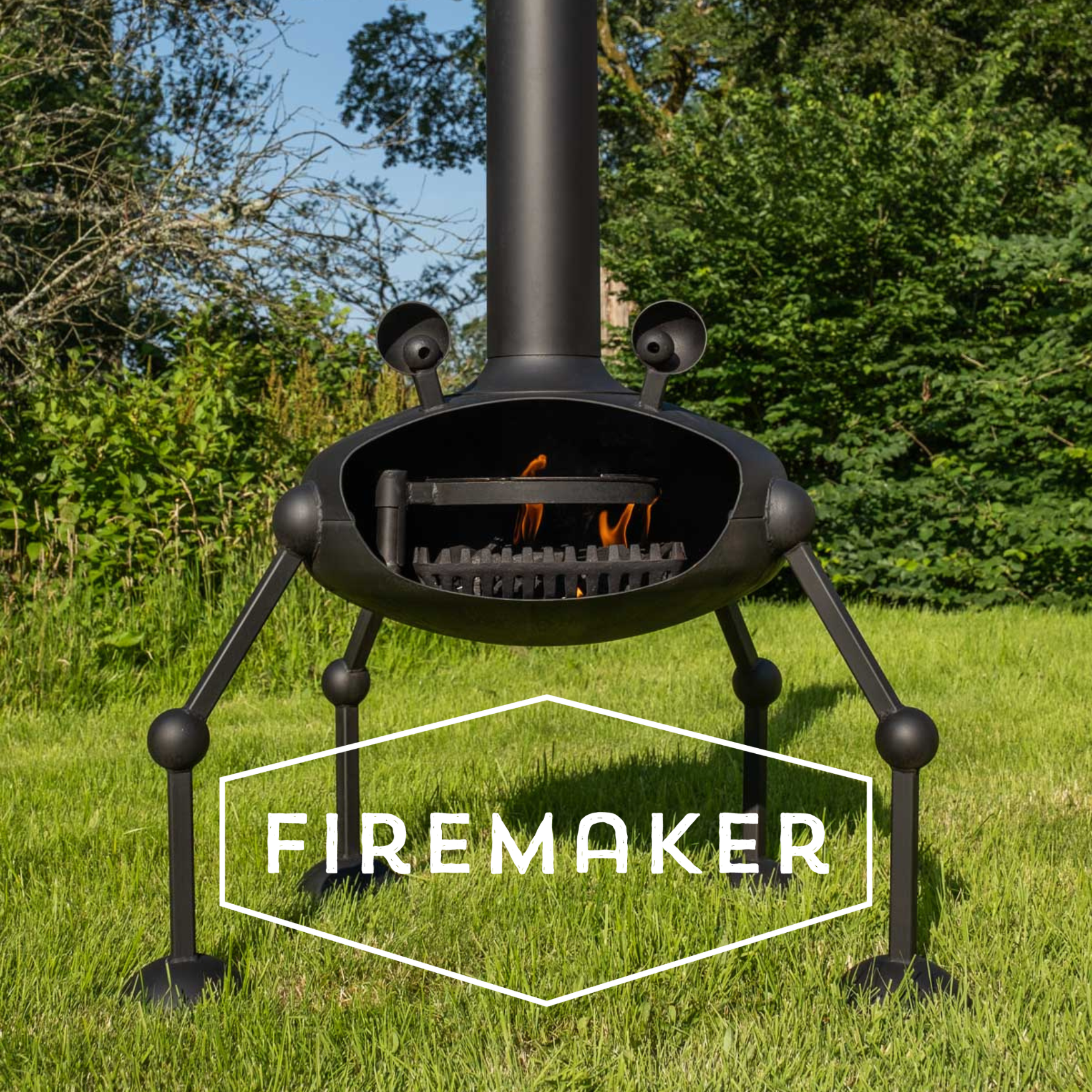 A black metal outdoor fire pit with a cylindrical chimney, and four legs, shown on a grassy lawn with trees in the background. A white hexagonal logo overlay at the bottom reads FIREMAKER.