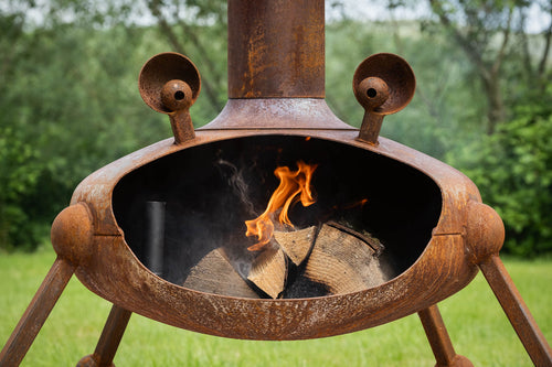A rustic metal campfire stove with a visible fire and logs inside, photographed outdoors on grass with trees in the background.