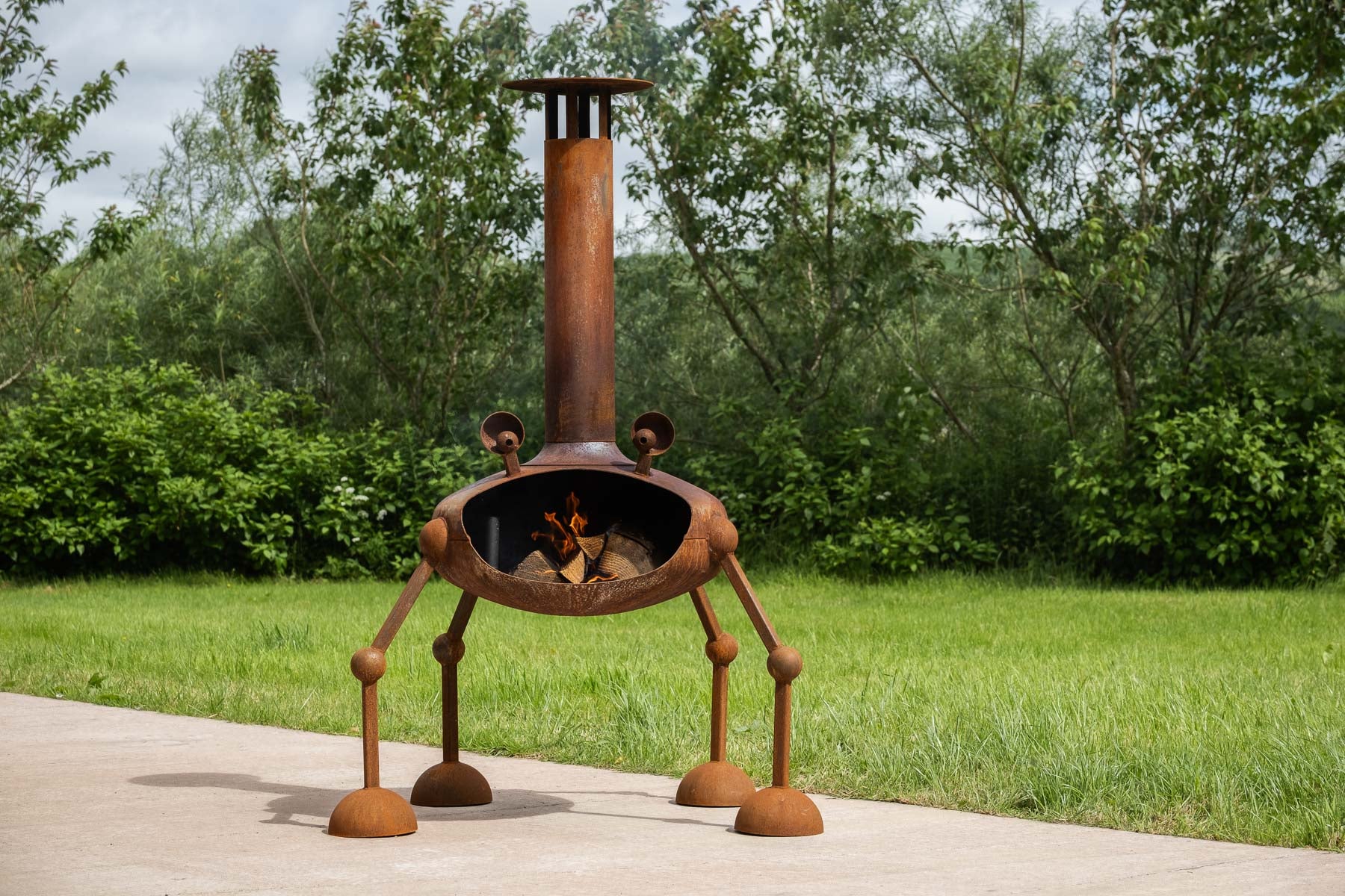 A rusted metal outdoor fire pit with four legs and a tall chimney, shown lit on a concrete patio with a green lawn and trees in the background.