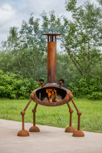 A rusted metal outdoor chiminea with four legs and a tall chimney, shown lit with visible flames, set on a concrete patio with a green lawn and trees in the background.