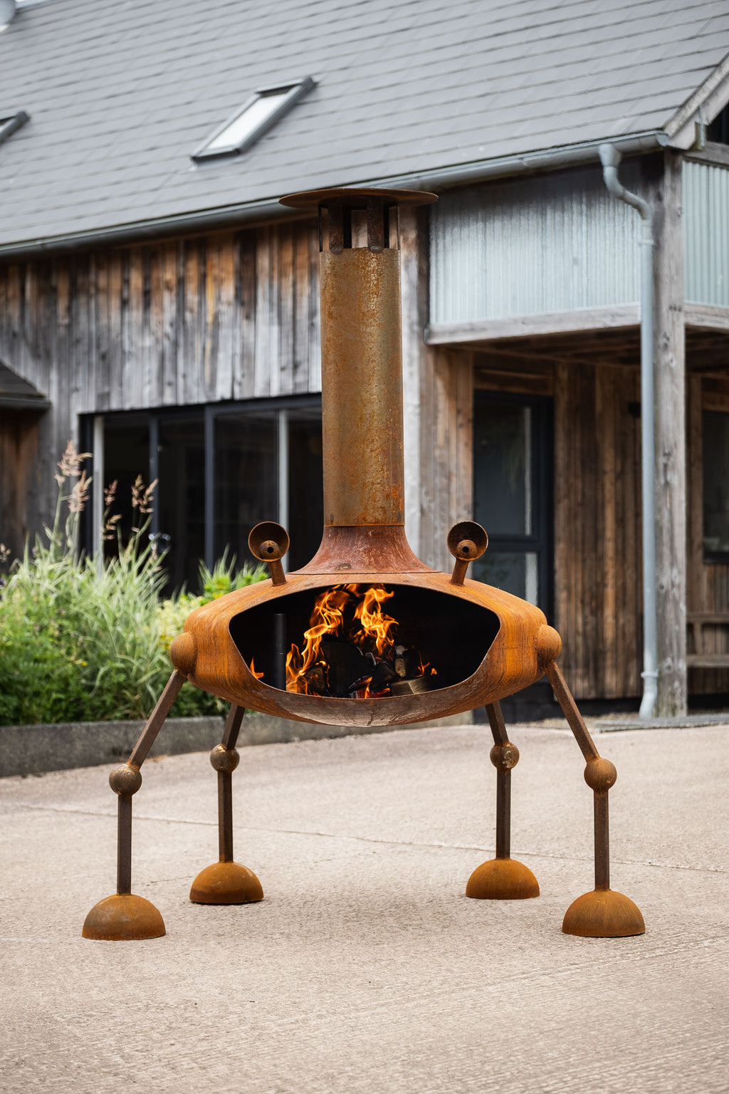 A rusted metal outdoor fire pit with a chimney and four legs, shown lit with flames in front of a rustic wooden building.