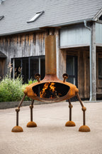 A rusted metal outdoor fire pit with a chimney and four legs, shown lit with flames in front of a rustic wooden building.
