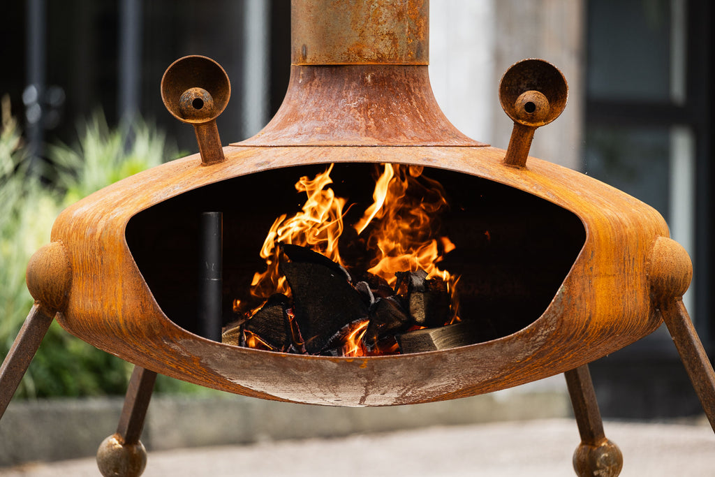 A rusted metal outdoor fire pit with a tri-legged base, open center, and visible flames burning inside, photographed outdoors with blurred background elements.