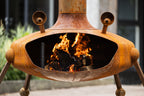 A rusted metal outdoor fire pit with a tri-legged base, open center, and visible flames burning inside, photographed outdoors with blurred background elements.
