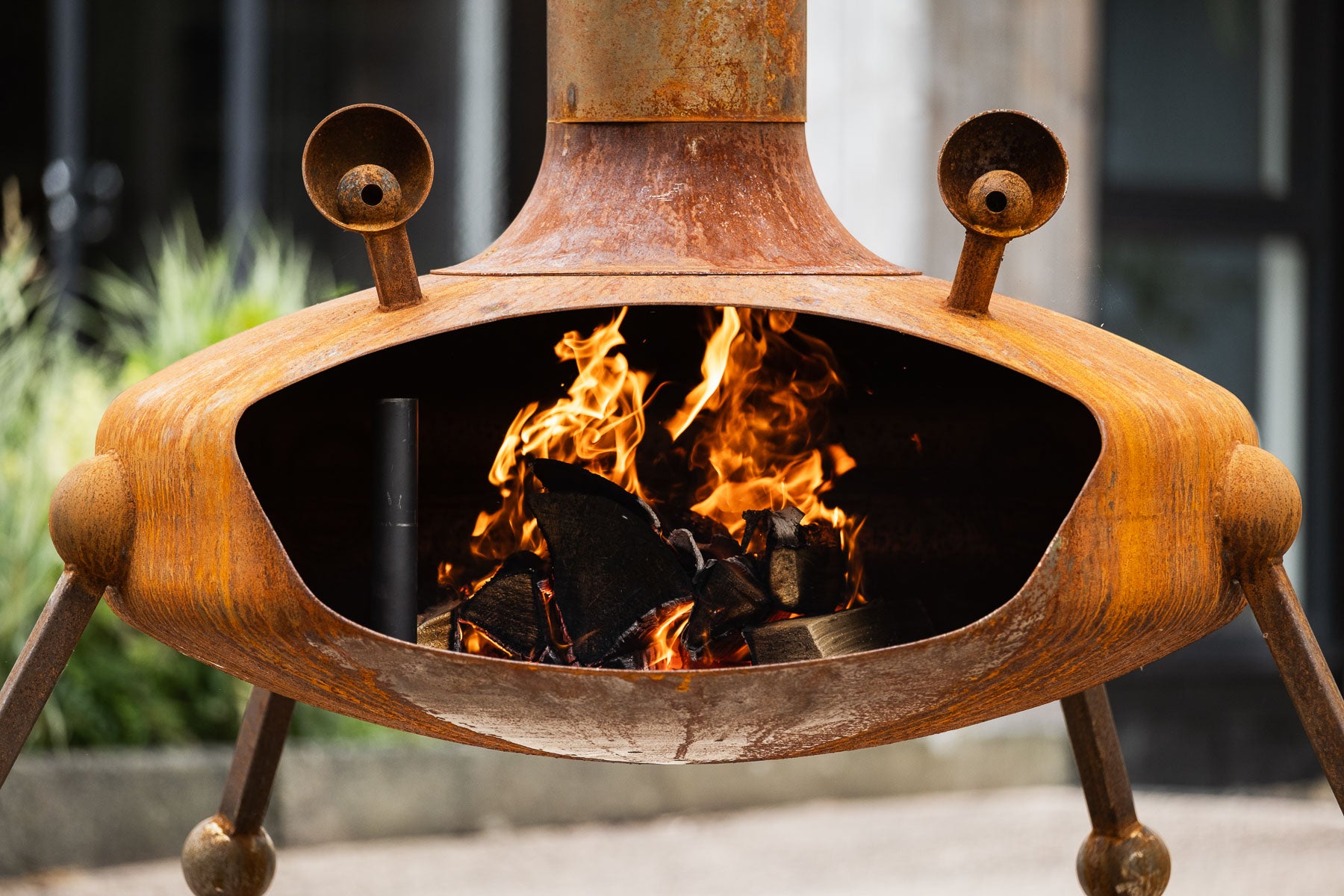 A rusted metal outdoor fire pit with a tri-legged base, open center, and visible flames burning inside, photographed outdoors with blurred background elements.