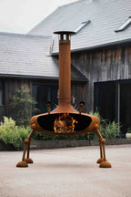 A rusted orange metal outdoor fire pit with a unique four-legged design and a tall chimney, shown lit with a fire inside, set on a concrete patio in front of a rustic wooden house.