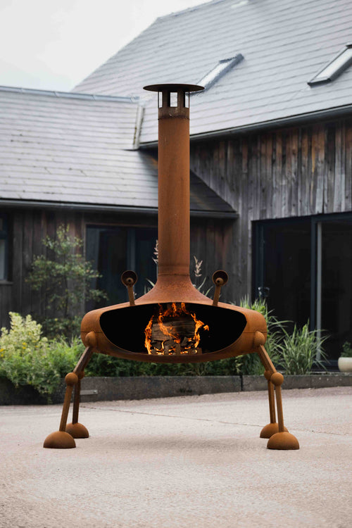 A rusted orange metal outdoor fire pit with a unique four-legged design and a tall chimney, shown lit with a fire inside, set on a concrete patio in front of a rustic wooden house.