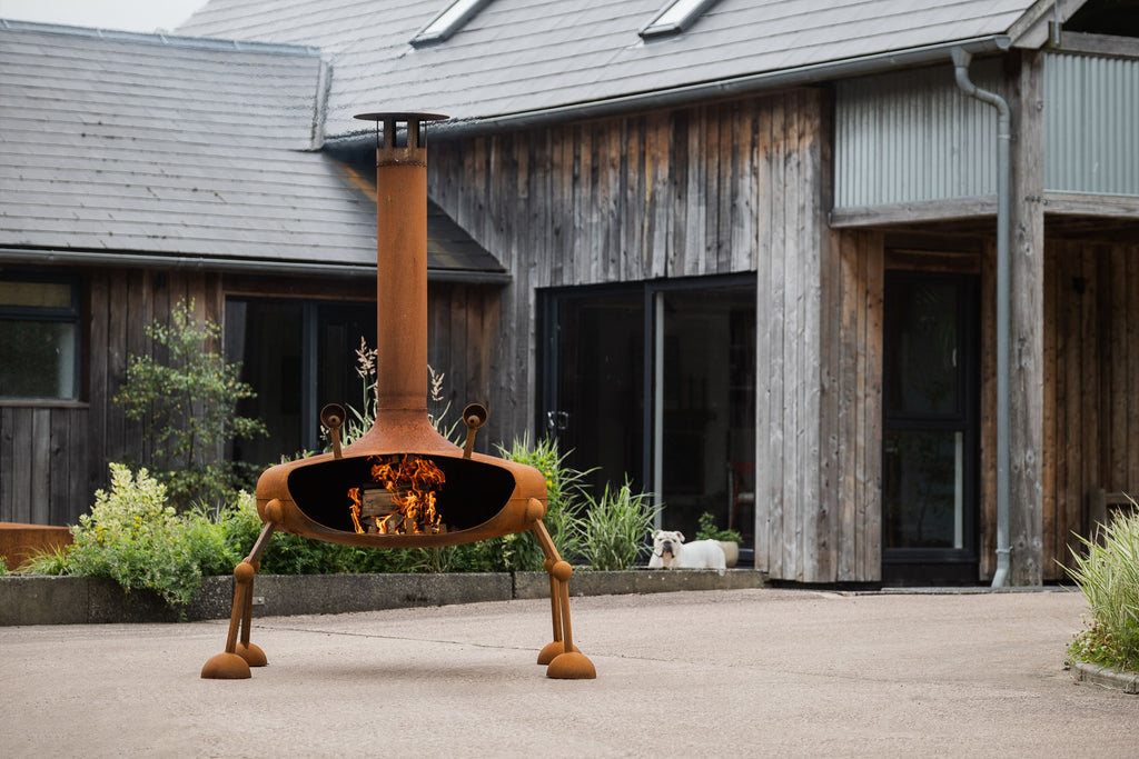 A rusted orange metal patio fire pit with four legs and a tall chimney, shown lit with flames in the center of a paved courtyard in front of a rustic wooden house.