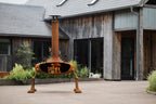 A rusted orange metal patio fire pit with four legs and a tall chimney, shown lit with flames in the center of a paved courtyard in front of a rustic wooden house.
