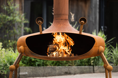 A rusted metal outdoor fire pit with a chimney and legs, shown lit with visible flames inside, set against a blurred garden background.