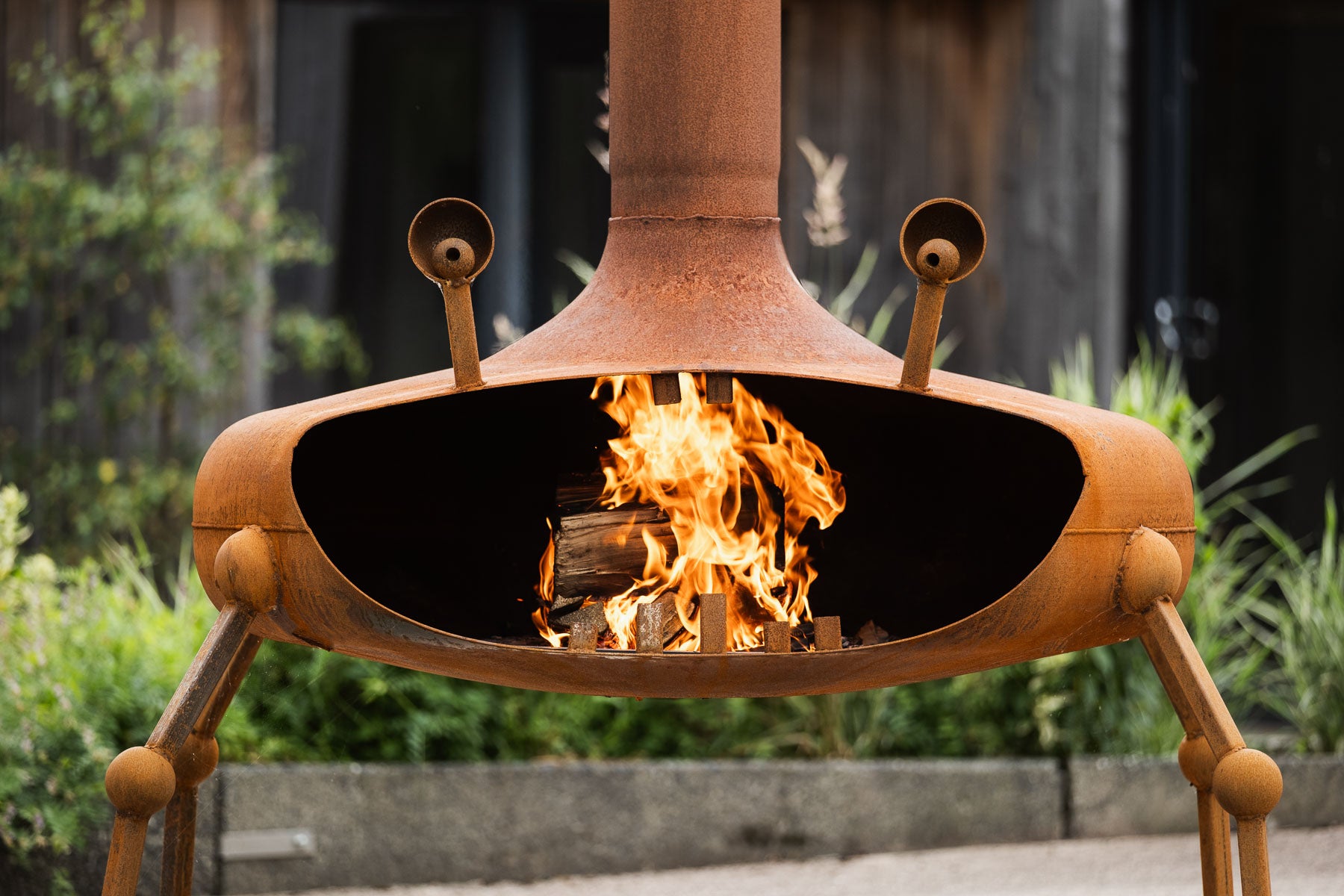 A rusted metal outdoor fire pit with a chimney and legs, shown lit with visible flames inside, set against a blurred garden background.
