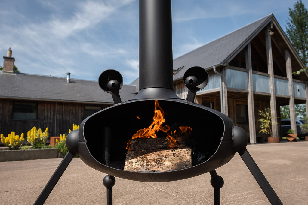 A black metal outdoor fire pit with a chimney and tripod legs, shown lit with a wood log burning inside, set in a backyard courtyard with a house and trees in the background.