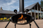 A black metal outdoor fire pit with a chimney and tripod legs, shown lit with a wood log burning inside, set in a backyard courtyard with a house and trees in the background.