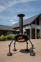 A black metal outdoor fireplace with a tall chimney and four legs, shown with a fire burning inside in a patio setting with a rustic cabin in the background.