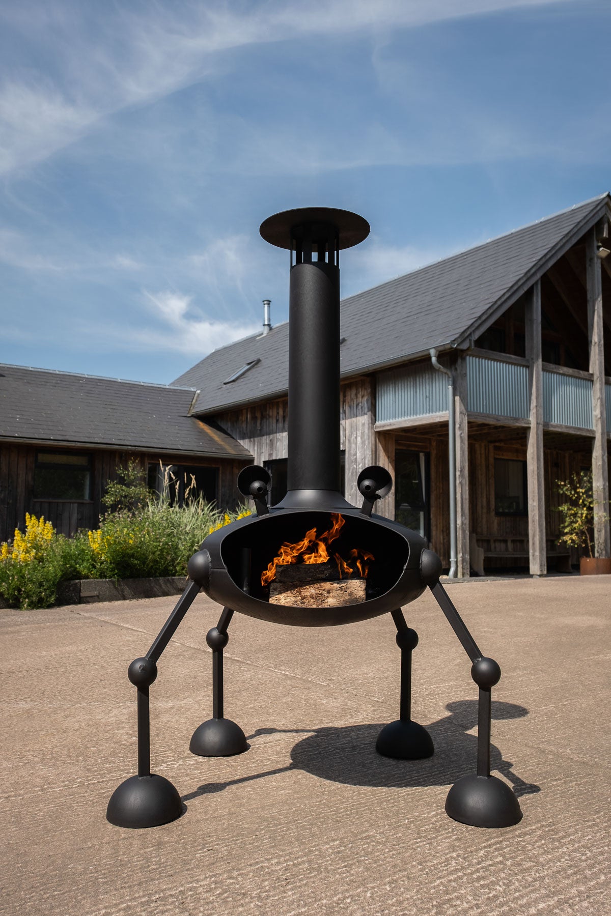 A black metal outdoor fireplace with a tall chimney and four legs, shown with a fire burning inside in a patio setting with a rustic cabin in the background.