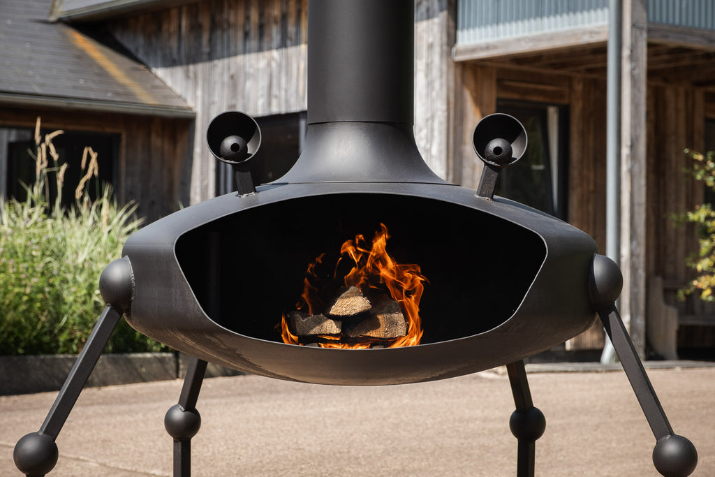 A black metal outdoor fire pit with a visible fire inside, shown on a patio in front of rustic wooden buildings.