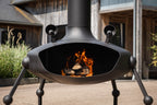 A black metal outdoor fire pit with a visible fire inside, shown on a patio in front of rustic wooden buildings.