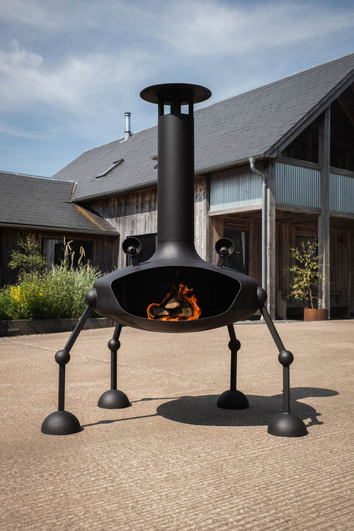 A modern black outdoor churrera fire pit with a tall chimney and four legs, shown lit with orange flames on a paved patio in front of a rustic wooden house.