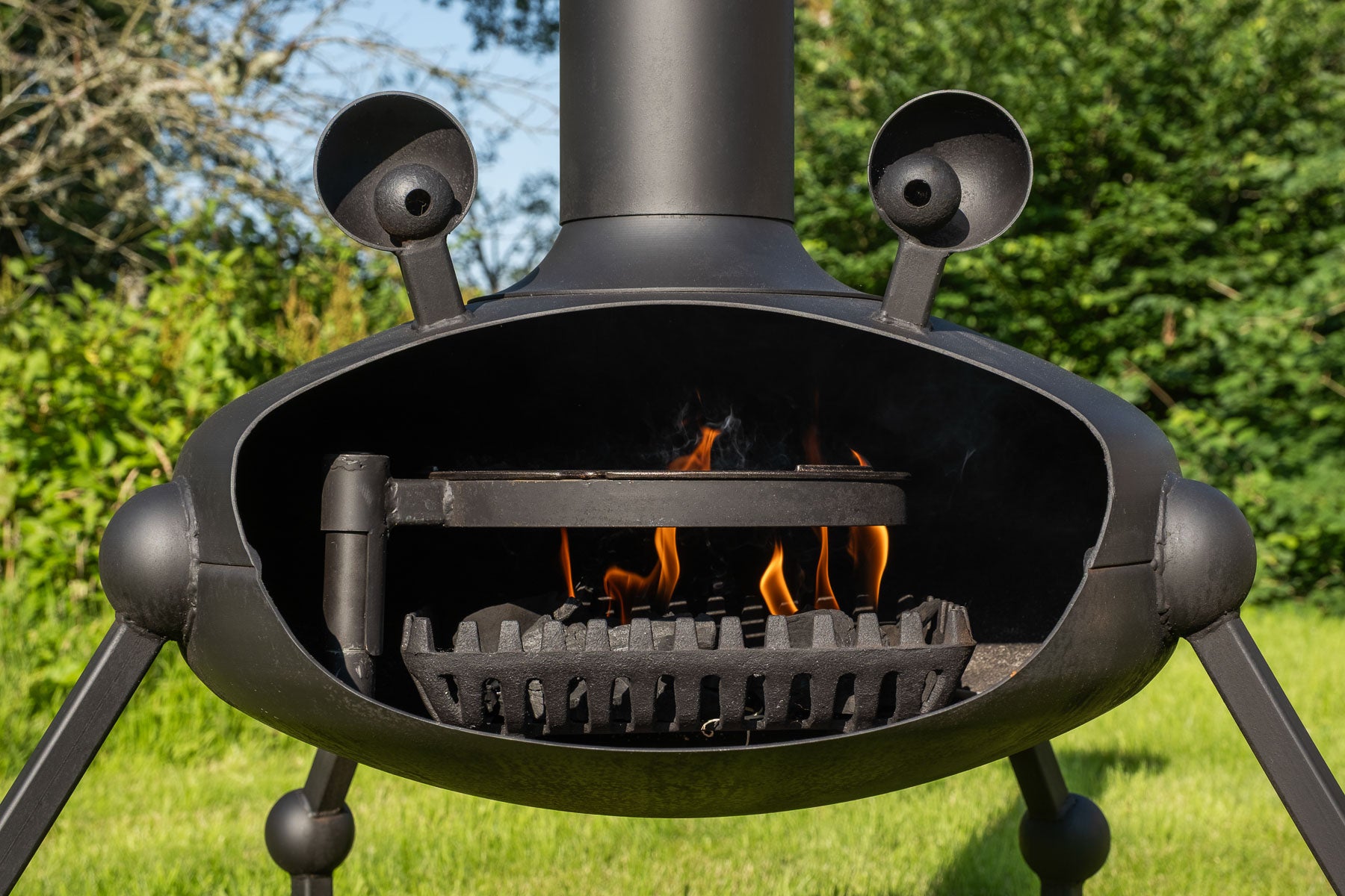 A black metal outdoor fire pit with a cylindrical chimney, swing grill and charcoal tray, and four legs, shown open with flames burning inside on a grassy lawn with trees in the background.
