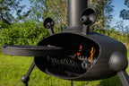 A black metal outdoor fire pit with a cylindrical chimney, swing grill extension, and two horn-like vents, shown lit with visible flames in an outdoor grassy setting.