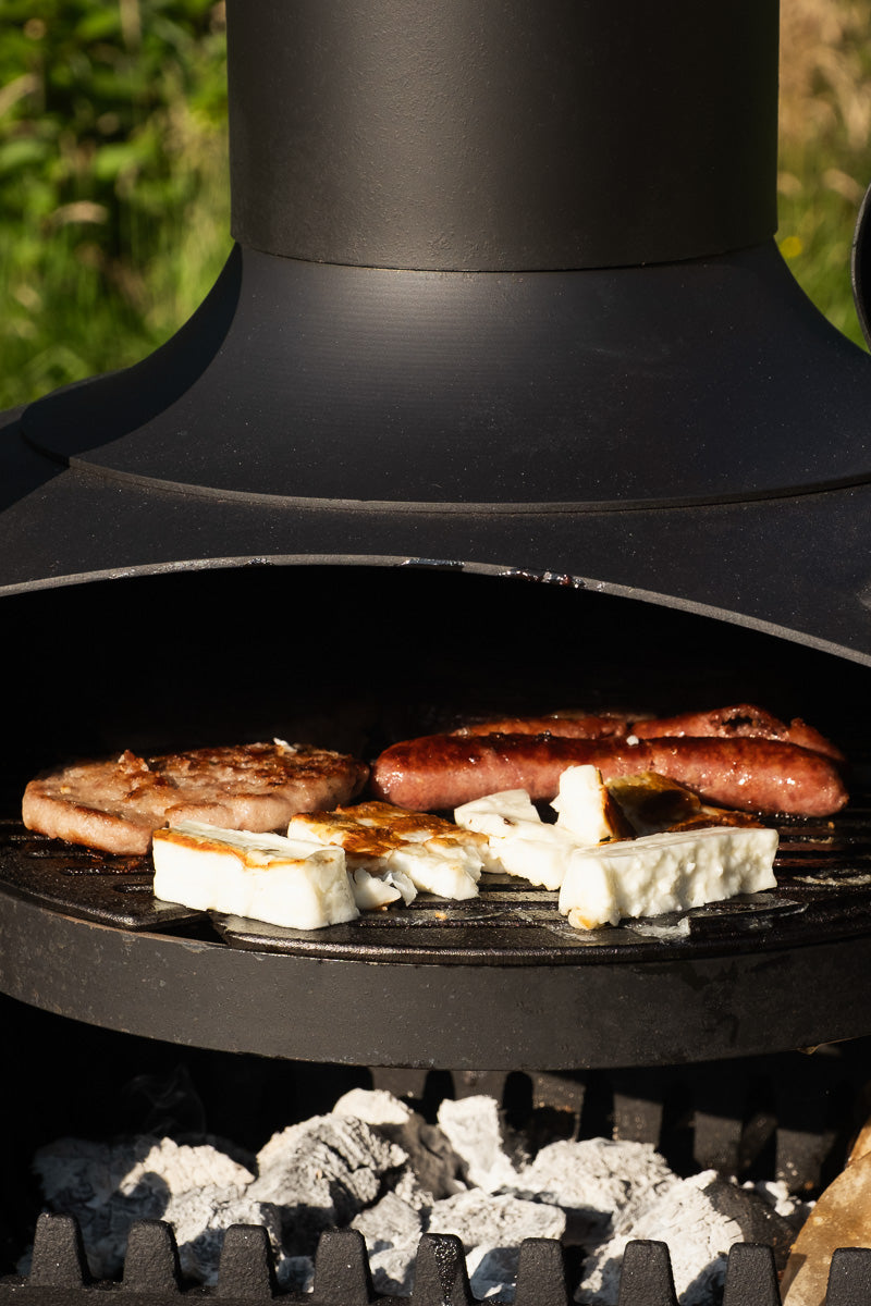A close-up outdoor shot of a black freestanding grill or smoker with food cooking on the grate, including sausages, a steak, and white cheese slices, with visible coals below.